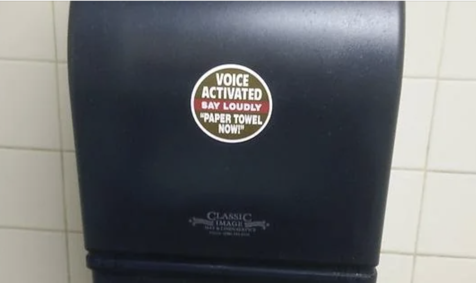 Paper towel dispenser with a sticker that reads, "VOICE ACTIVATED. SAY LOUDLY 'PAPER TOWEL NOW!'"