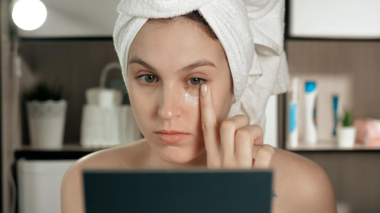 Eye cream. Young attractive woman applies cream to skin under her left eye, girl is in bathroom and looks in mirror. Hygiene, beautician, makeup, face care concept. Close-up view