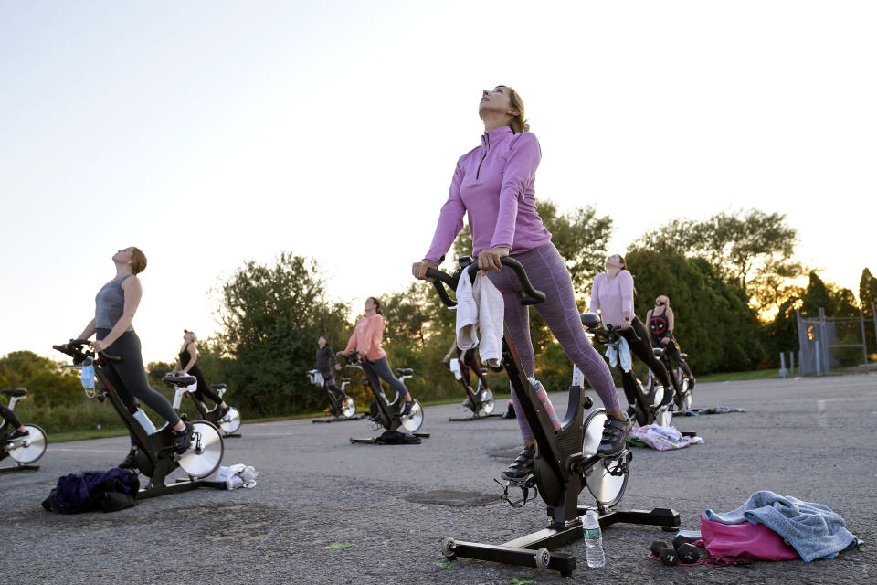 Donna Coskren, of Newburyport, Mass., front, stretches with others while on stationary exercise bikes during a spinning class in a parking lot outside Fuel Training Studio, Monday, Sept. 21, 2020, in Newburyport. The gym's revenue is down about 60% during the COVID-19 pandemic. Fuel Training Studio plans to continue holding outdoor classes into the winter with the help of a planned greenhouse-like structure with heaters but no walls. (AP Photo/Steven Senne)