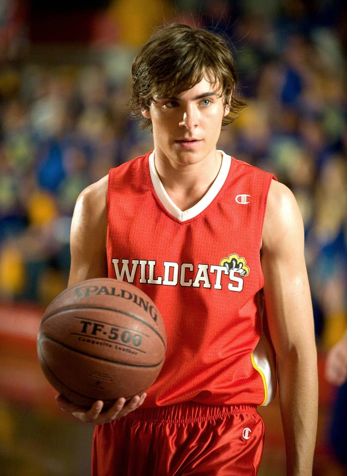 Zac Efron in a 'Wildcats' basketball uniform holding a basketball, standing on a court in a scene from High School Musical