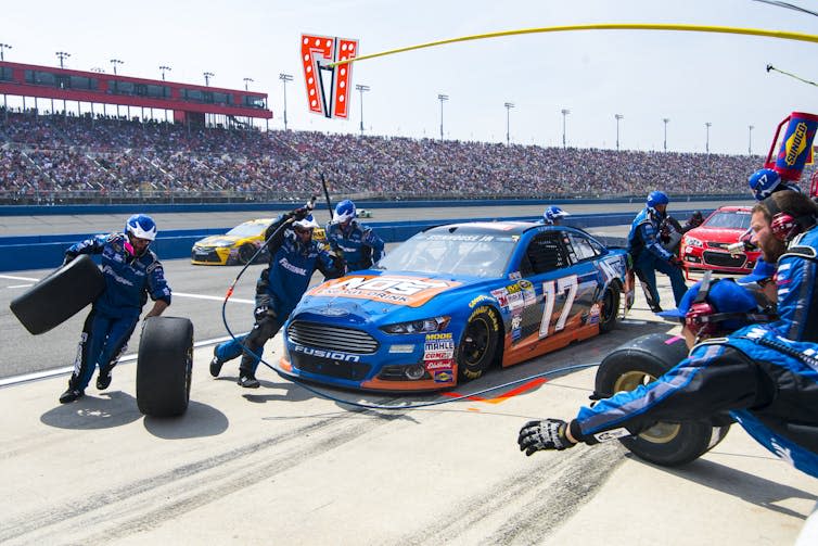 A Ford race car comes in for service during a race in front of a packed grandstand.