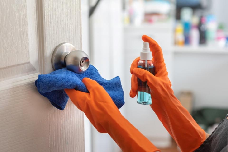 Person wearing orange rubber gloves disinfecting door knob using spray and a blue cloth.