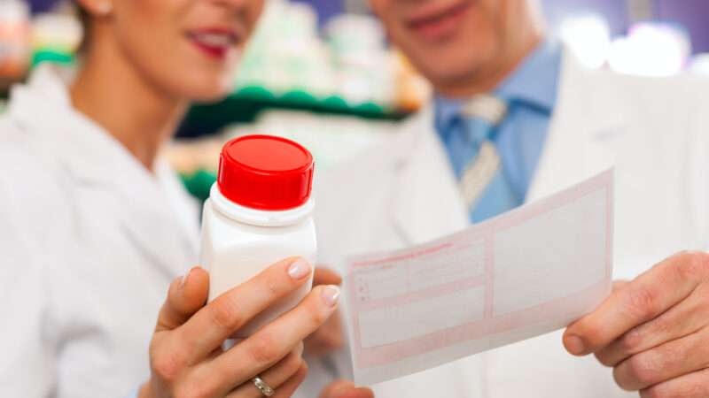 Two pharmacists consult one another, one holding a pill bottle and the other holding a prescription.