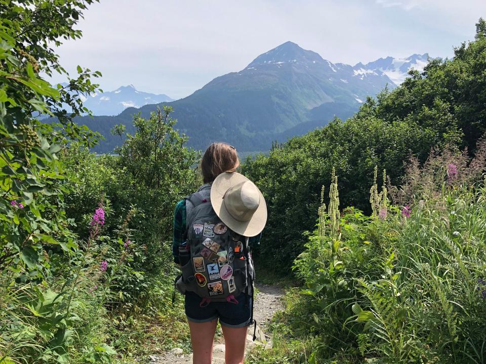 Emily, wearing a backpack covered in patches and a sunhat on her back, hikes through a grassy trail in Kenai Fjords National Park.