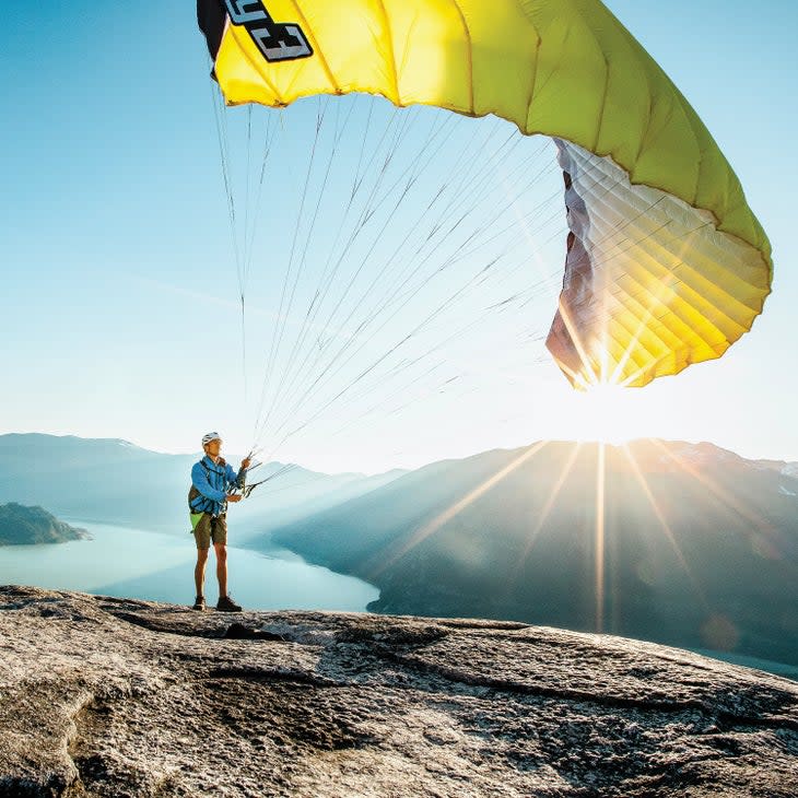 Earle about to go paragliding. He's standing on a flat dome mountain top. His unfurling glider wing is yellow. Mountains and a bay and the rising sun behind him.