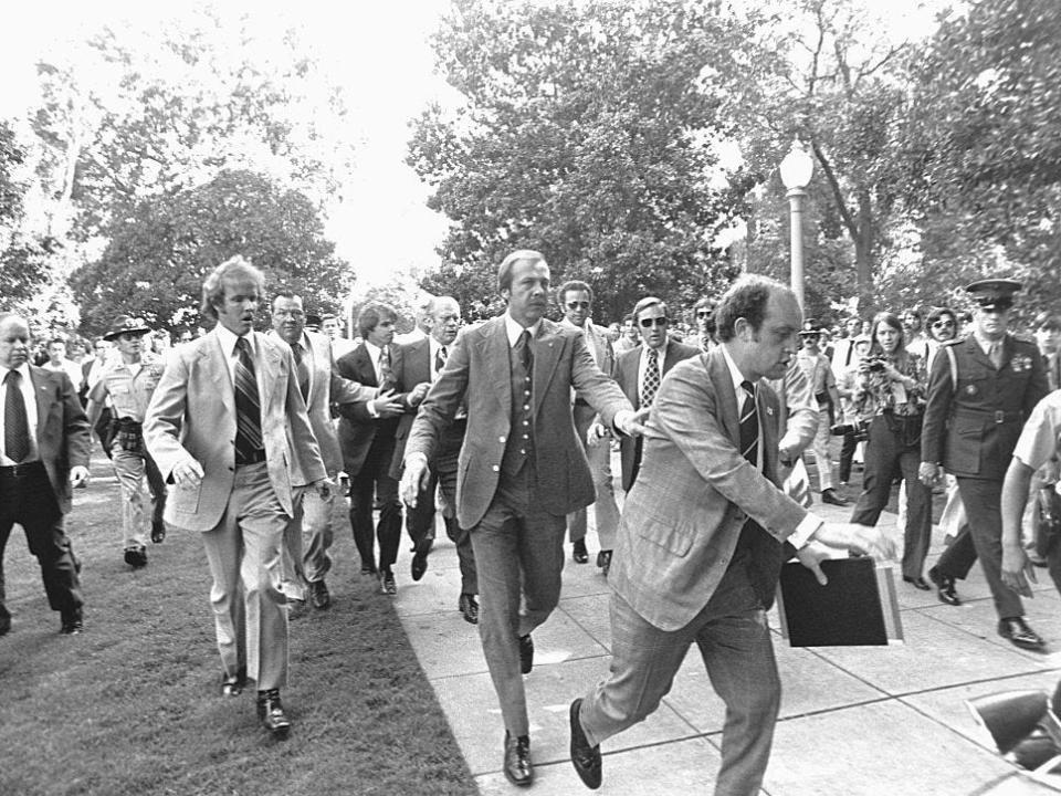 Police and secret service officers run to protect President Gerald Ford during an assassination attempt.