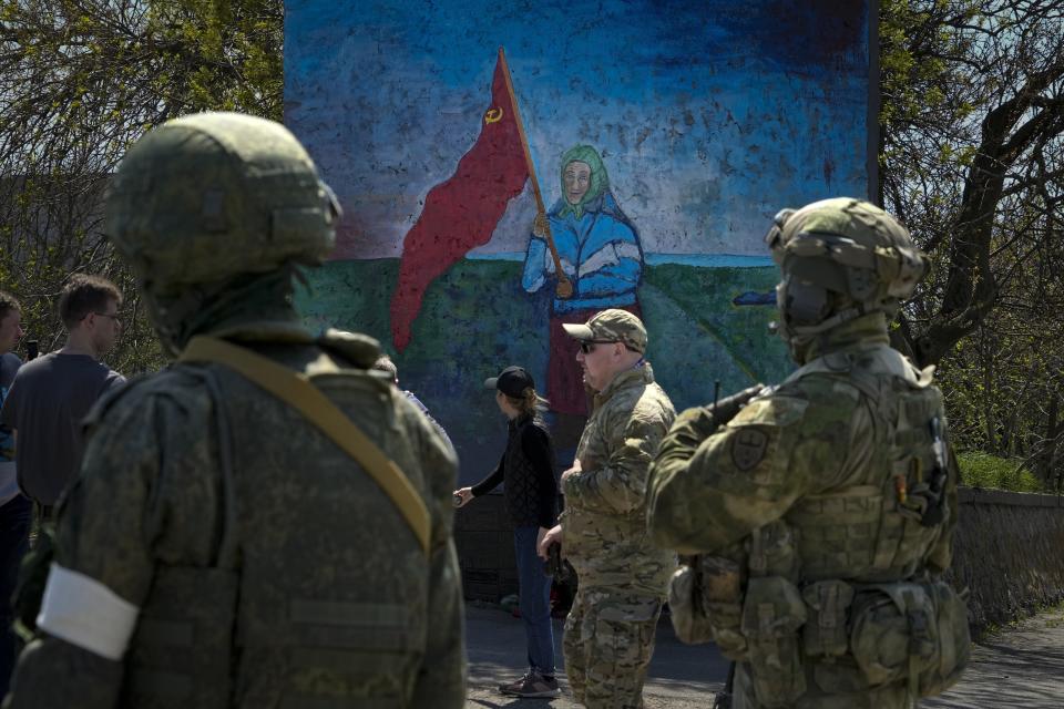 FILE - Russian servicemen stand near a painting of a Ukrainian woman holding a Soviet-era red flag in territory under the government of the Donetsk People's Republic, eastern Ukraine, April 30, 2022. Some in the West think Russian President Vladimir Putin may use the Victory Day on May 9 when Russia celebrates the defeat of Nazi Germany in World War II to officially declare that war is underway in Ukraine and announce a mobilization _ the claim rejected by the Kremlin. (AP Photo/Alexander Zemlianichenko, File)
