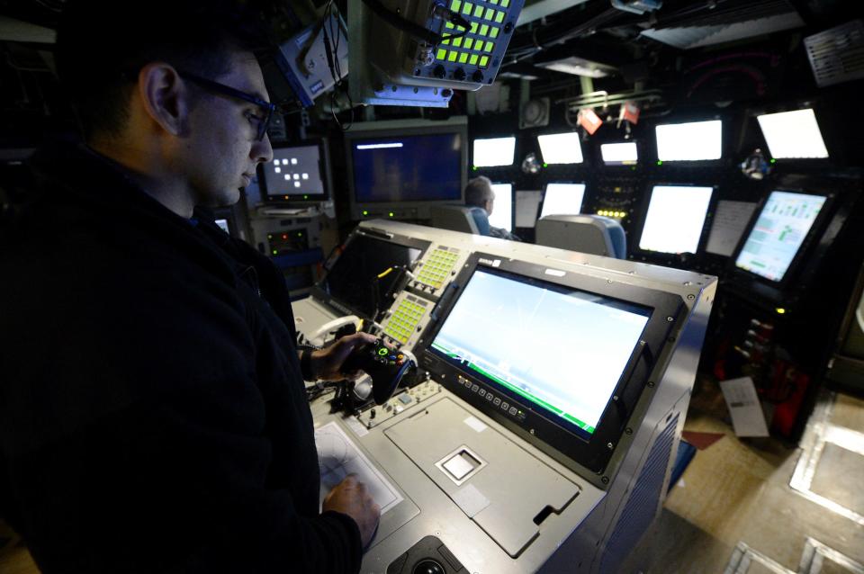 A Navy sailor in front of several computer monitors uses an xbox controller for periscope on submarine