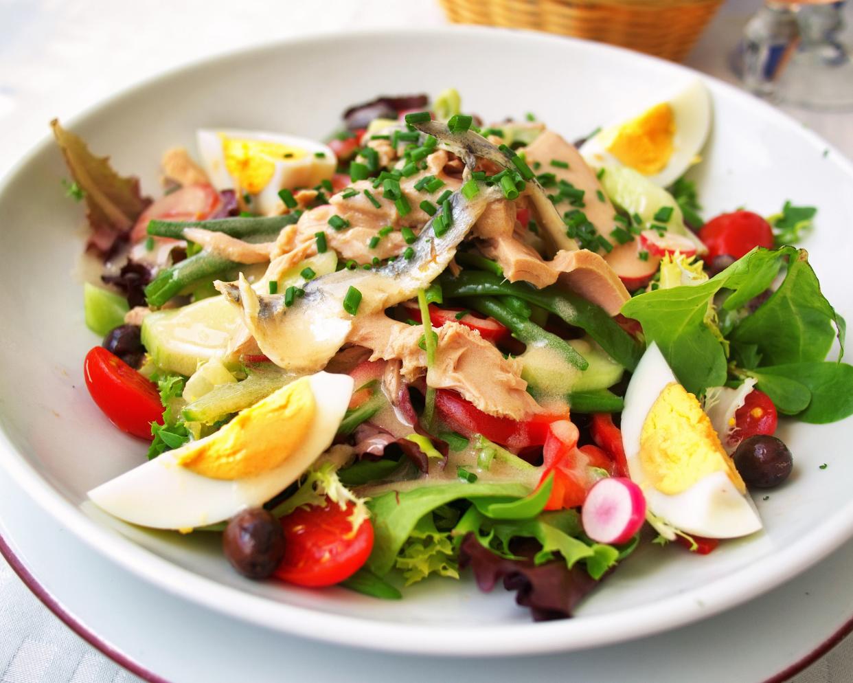 Nicoise salad in a shallow round white porcelain bowl, selective focus, on a purple rimmed white porcelain plate on a table with a basket blurred in the background