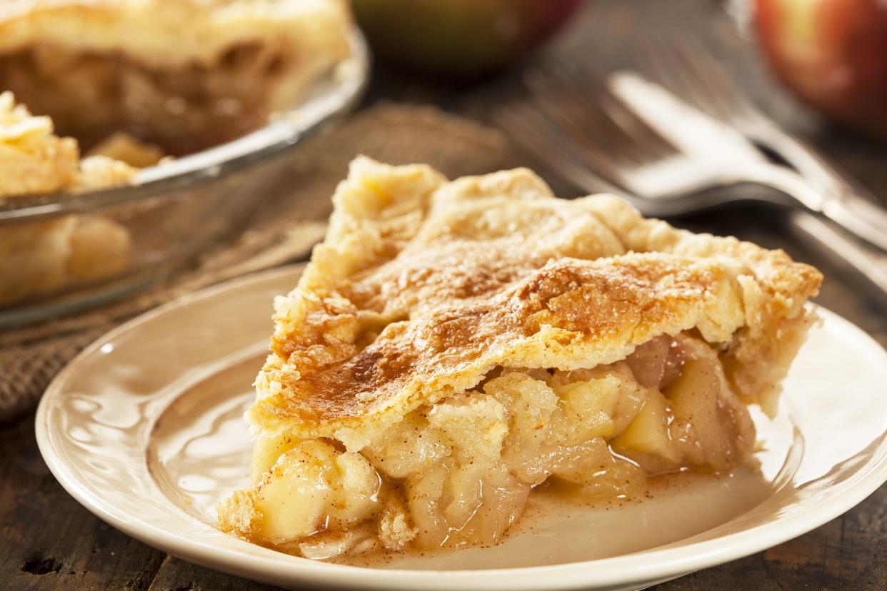 Sugar-free apple pie on a white plate with blurred background of rest of pie, utensils, and apples
