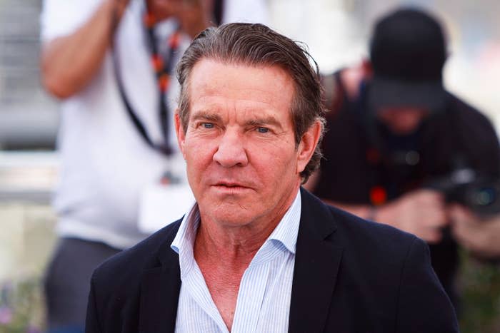 Dennis Quaid wearing a casual blazer over a collared shirt, standing at an event with a blurred background