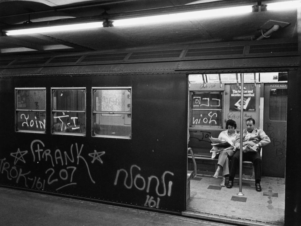 A subway car marked with extensive graffiti waits at a station platform, with the door open and two people seated inside