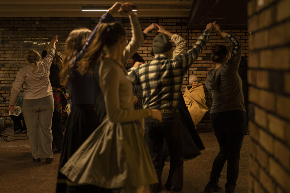 Una mujer mayor, cubierta por una manta, mira mientras varias personas bailan en una estación de metro luego de que sonaron las alarmas antiaéreas, en Kiev, Ucrania, el 1 de marzo de 2023. (AP Foto/Vadim Ghirda)