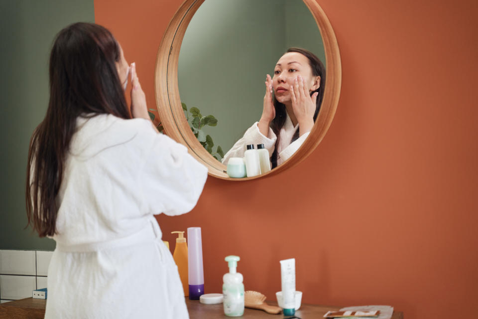 A dark-haired woman stands before a round mirror and applies skincare products. The mirror has a wooden frame and is hung on a terracotta coloured wall above a bench. The woman wears a white bathrobe.