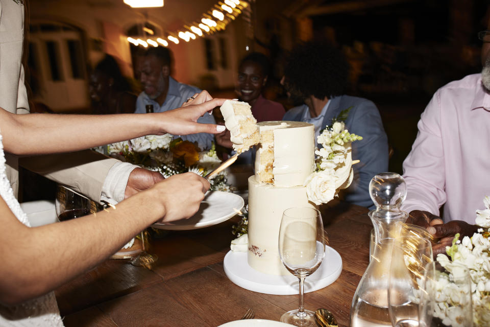 A person is cutting a slice of wedding cake on a table surrounded by people in formal attire at an evening event