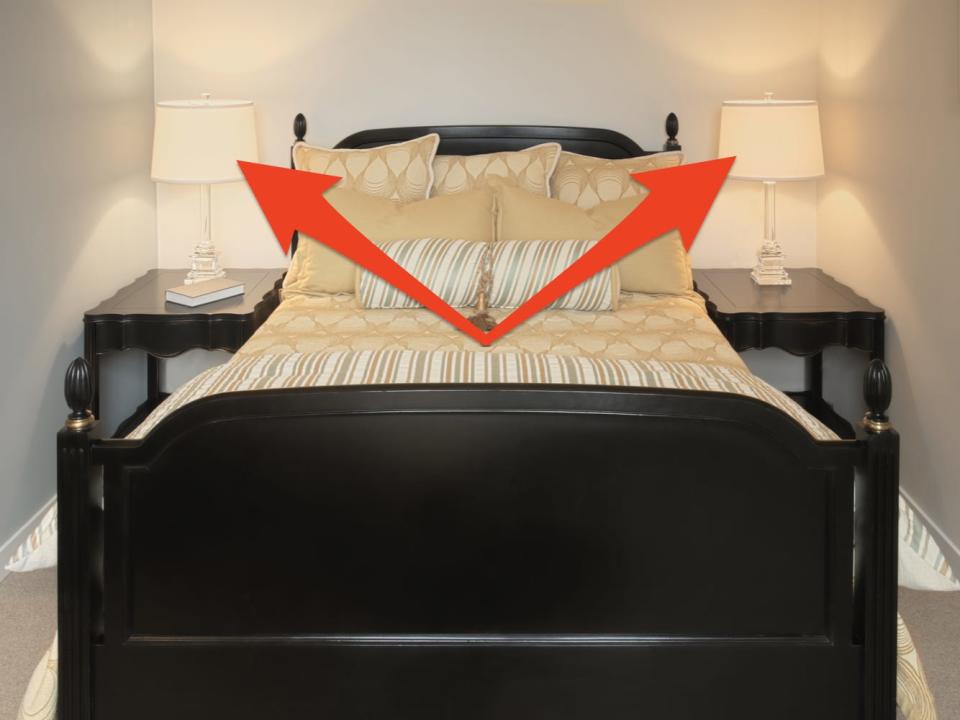 bed with black-painted wooden frame, golden sheets and pillows and matching black-painted nightstands with matching lamps on either side. Two red arrows point to the nightstands and lamps