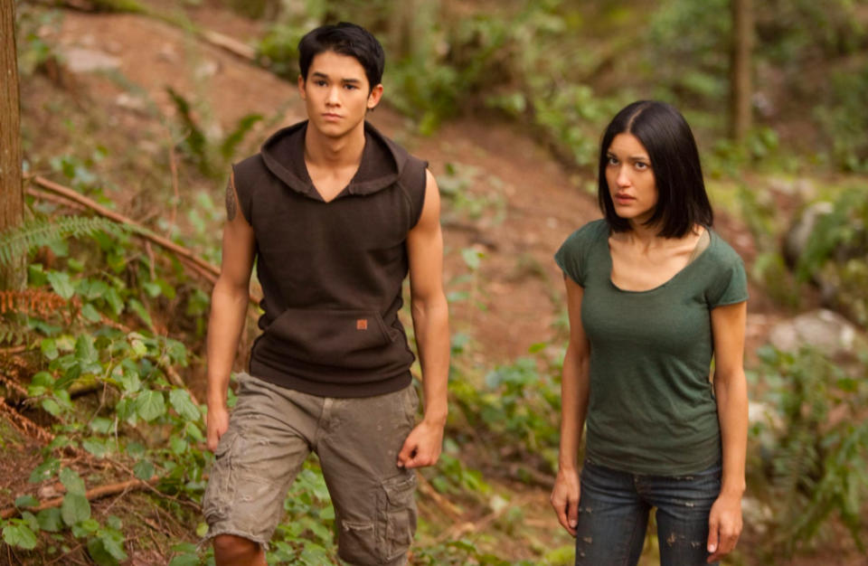 Booboo Stewart in a hooded sleeveless shirt and Tinsel Korey in a short-sleeve shirt stand in a forest. Both appear focused and intense