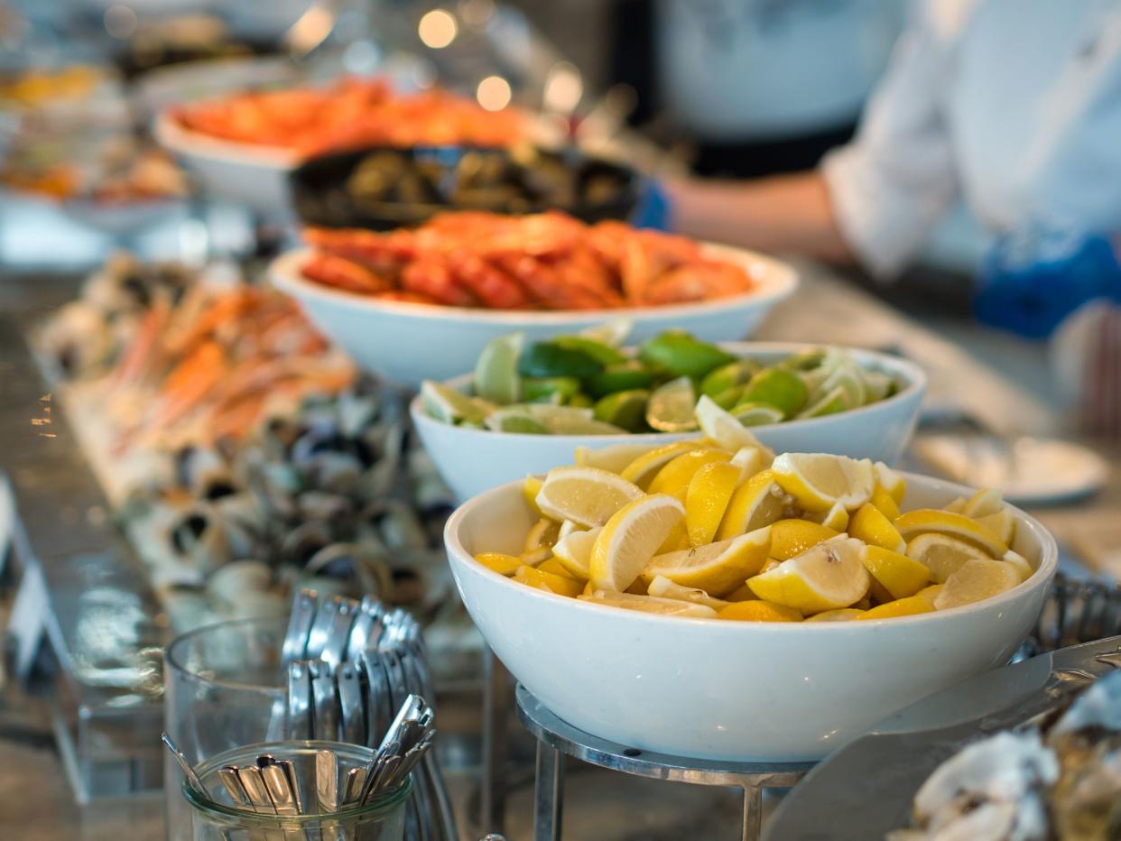 A large bowl of freshly sliced lemon hoisted on a stand along with a similarly large bowl of sliced lime and cold seafoods in a blurred background.