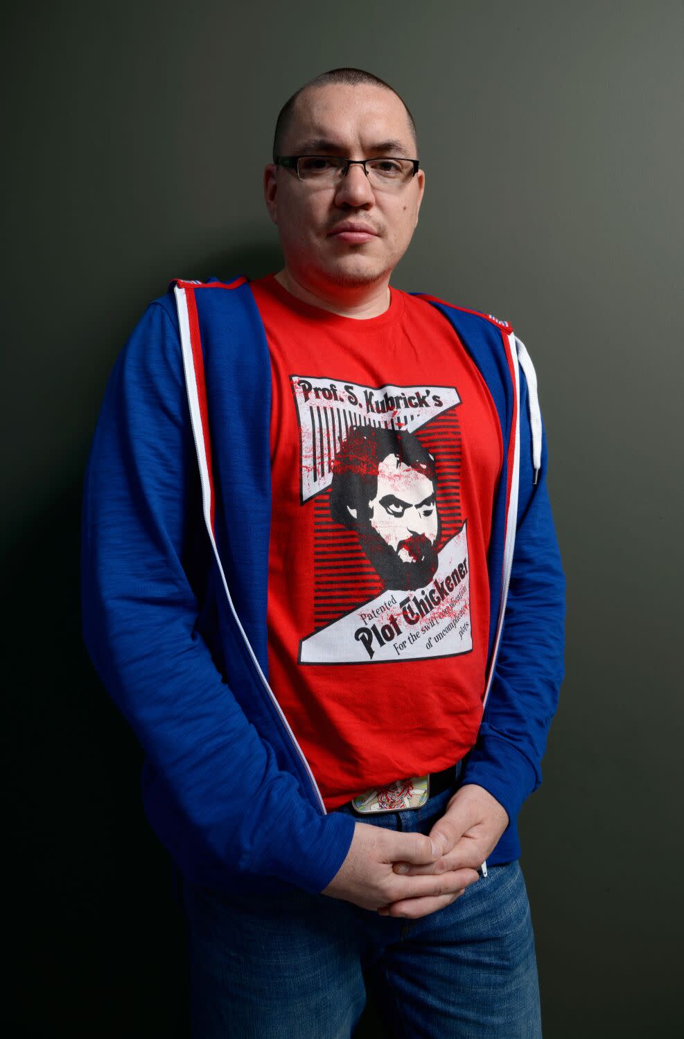 A man in a red T-shirt and blue hoodie stands with hands clasped, looking at the camera.
