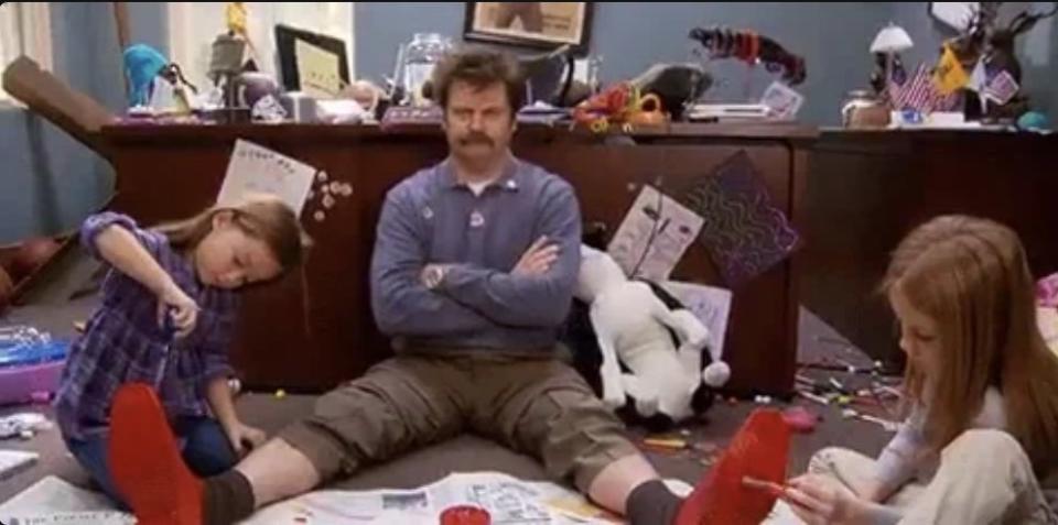 Man sits cross-legged on the floor, with two children painting his feet, surrounded by a cluttered room filled with papers, toys, and crafts