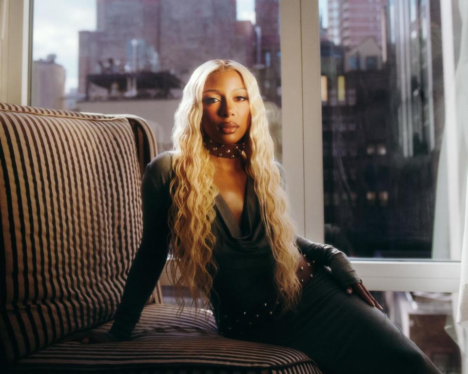 A woman with long blond hair sits on a couch, buildings visible through the window behind her