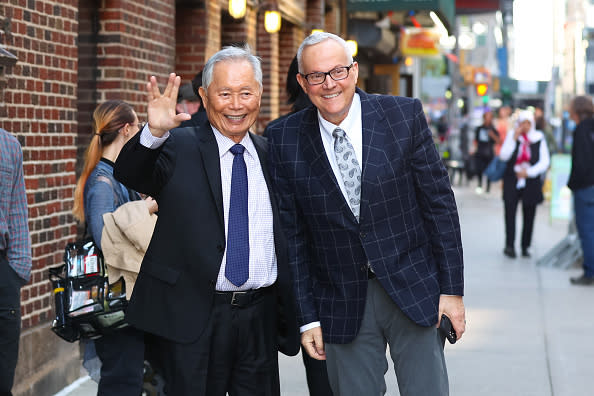 George Takei and Brad Altman smiling and posing together on a city sidewalk. George is waving. Brad is wearing a patterned blazer