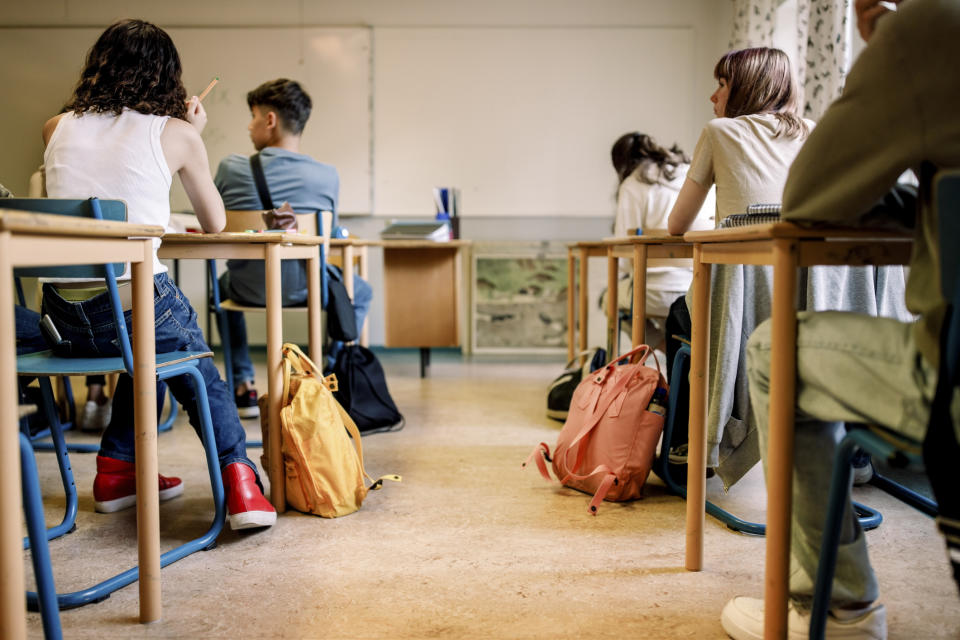 Students seated in a classroom facing the front, some with notebooks, next to backpacks