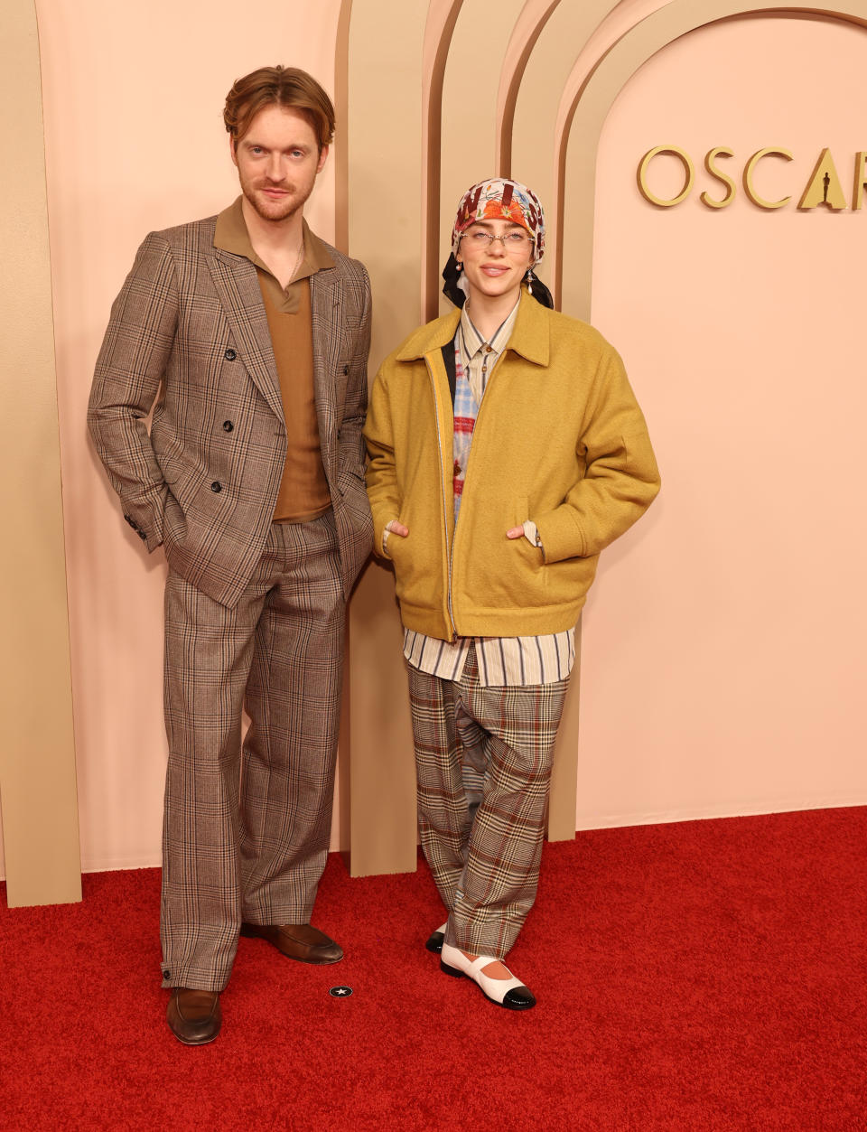 Finneas and Billie standing in front of an "OSCARS" backdrop. Finneas is wearing a checked suit
