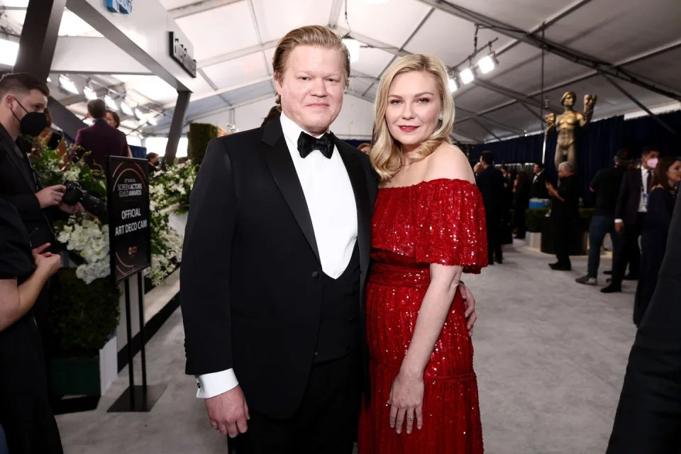 jesse plemons and kirsten dunst stand together embracing as they look at the camera and smile, he wears a black tuxedo with a matching bow tie and white collared shirt, she wears an off the shoulder red sparkling dress