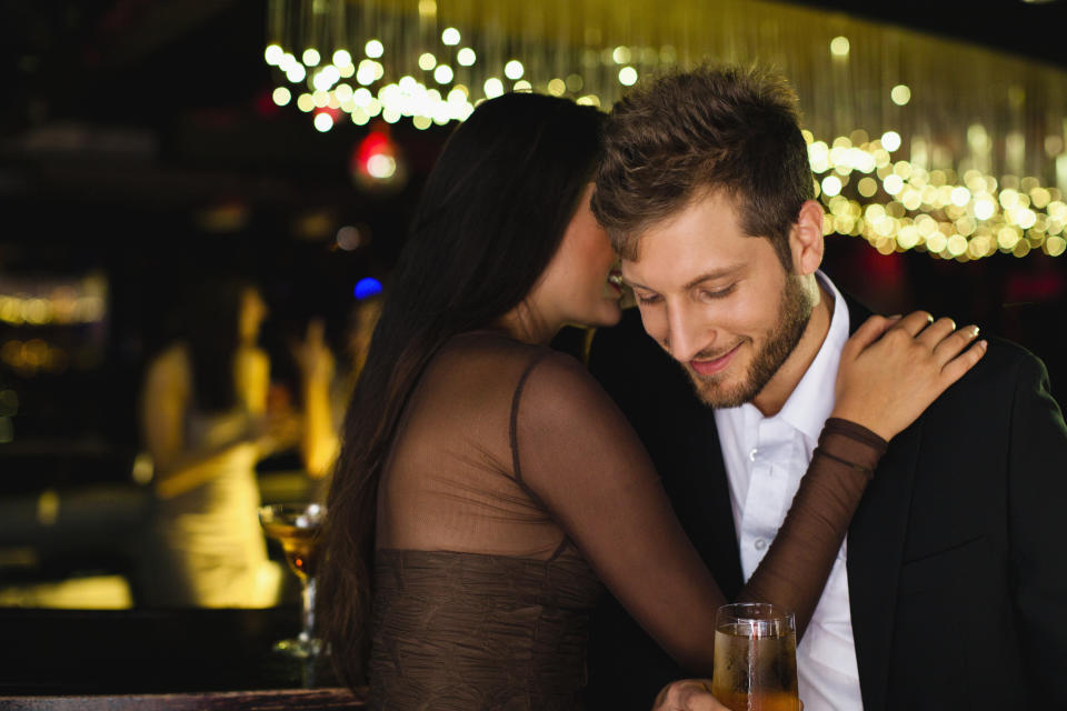 A woman and a man share an intimate moment at a dimly lit bar as she whispers in his ear. He smiles while holding a drink, and she has her arm around his shoulder