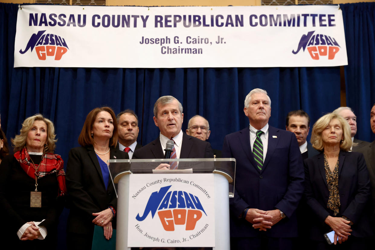 Nassau County Republican Party chairman Joseph Cairo at a podium surrounded by local GOP officials at a podium and under a banner both reading: Nassau County Republican Committee, Joseph G. Cairo, Jr, Chairman..