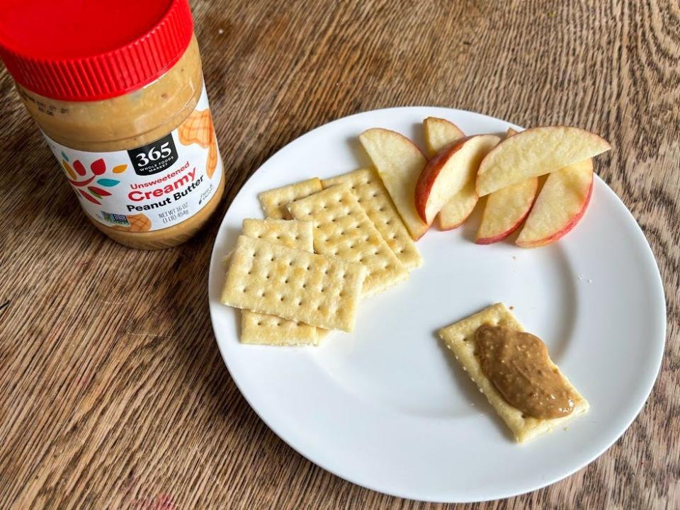 A jar of peanut butter with a white label and red lid next to a white plate of crackers and apple slices. A slightly chunky-looking peanut butter is spread on one cracker