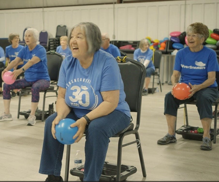 A group of older adults in blue T-shirts doing chair exercises as part of a SilverSneakers fitness class.