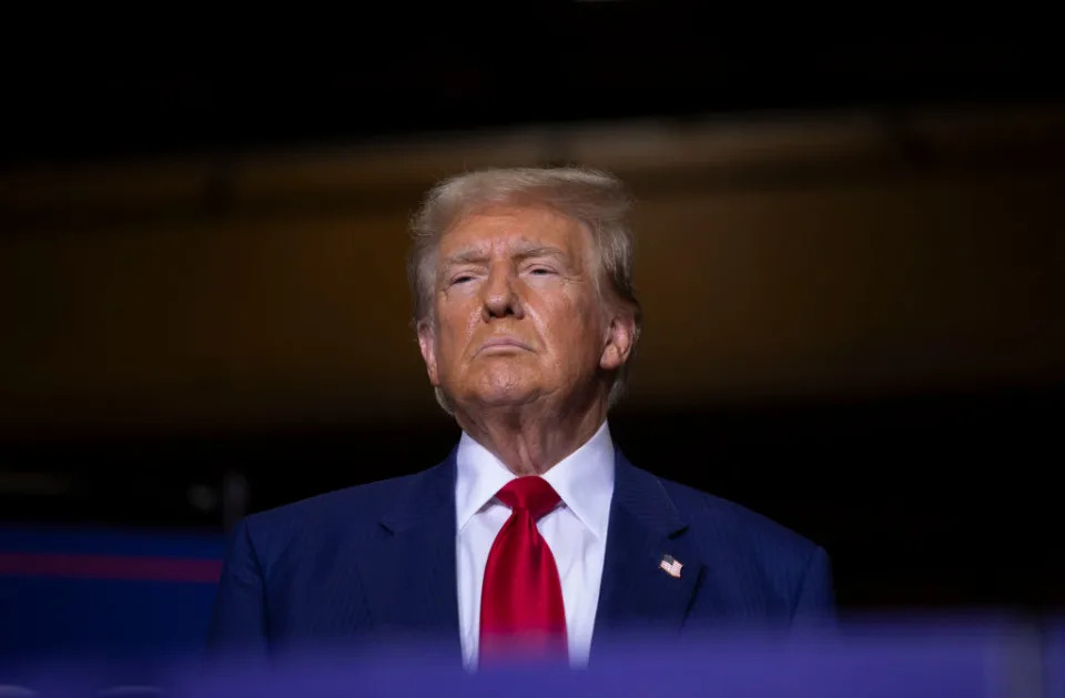 Donald Trump stands in a dark setting wearing a suit with a white shirt and red tie, looking upwards with a serious expression