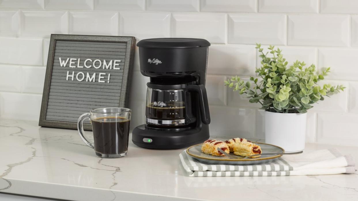  Mr. Coffee 5-Cup Mini Brew Coffee Maker on marble countertop with signage, pastries, dish towel and indoor houseplant. 
