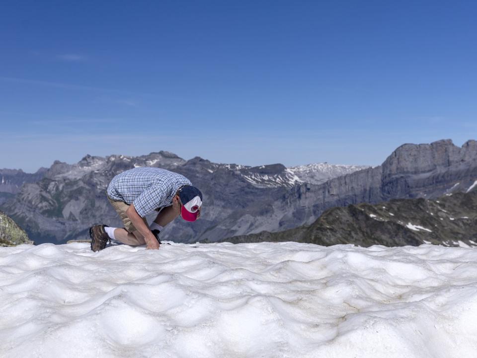 man in shorts button up and cap kneels in snow with mountains in the background