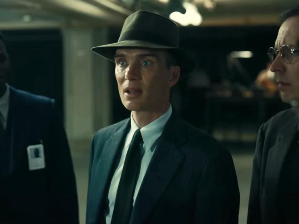 Cillian Murphy in a scene from "Oppenheimer" where J. Robert Oppenheimer is standing next to two men in suits while speaking.