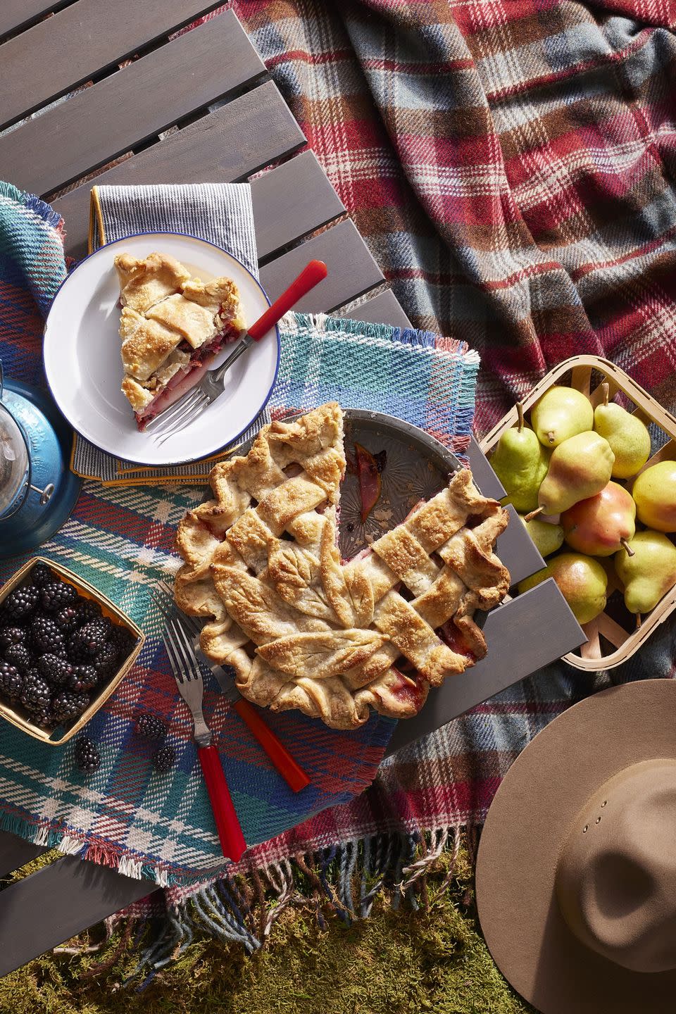 blackberry and pear pie with a lattice crust on top in a pie dish on a table outside with a slice on a white plate next to the pie and a small container of blackberries