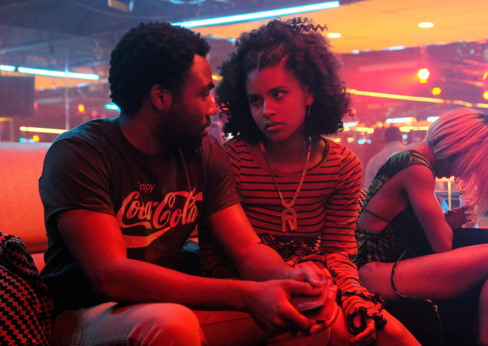 Donald Glover and Zazie Beetz sitting on a couch in a dimly lit club. Donald wears a casual T-shirt while Zazie is in a striped shirt