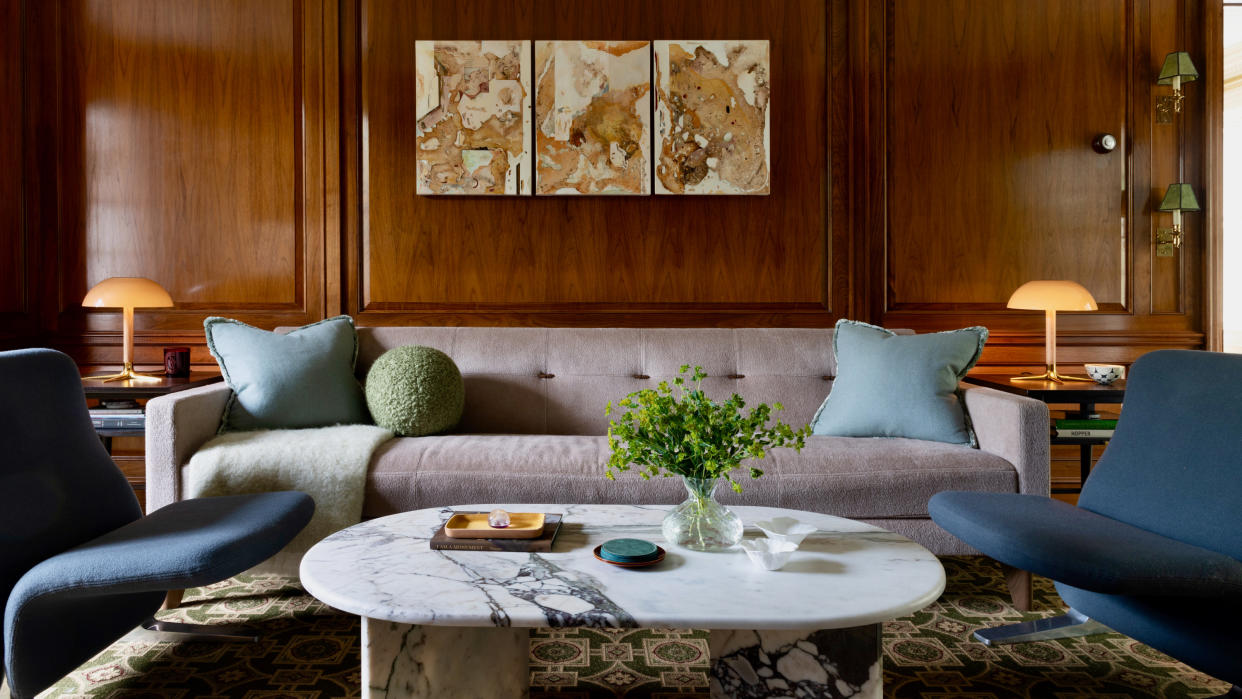  Living room with rich wood-panelled walls, lilac low-slung sofa and cool dark blue armchairs with marble coffee table. 