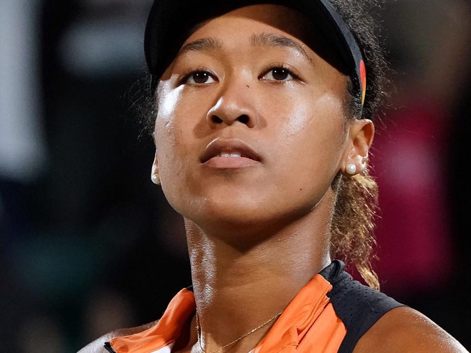 Naomi Osaka looks pensive while wearing a black and orange tennis shirt and visor and holding two tennis balls