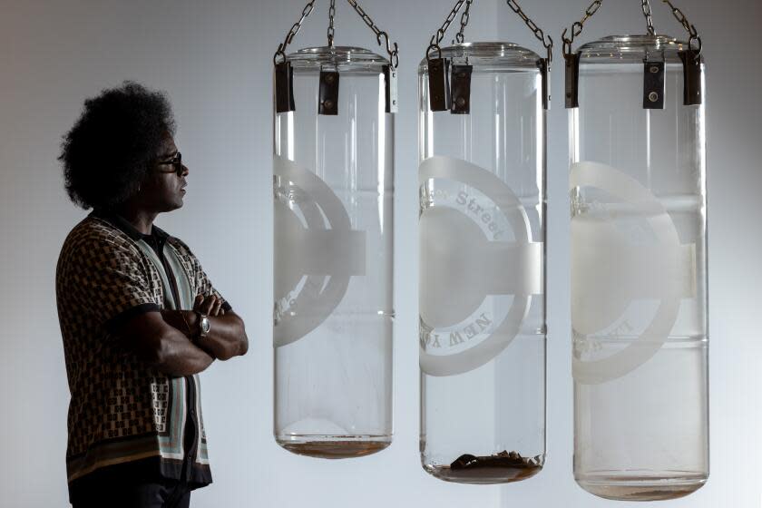 Alexandre Arrechea, a Black man in sunglasses wearing a geometric shirt, stands before three glass punching bags