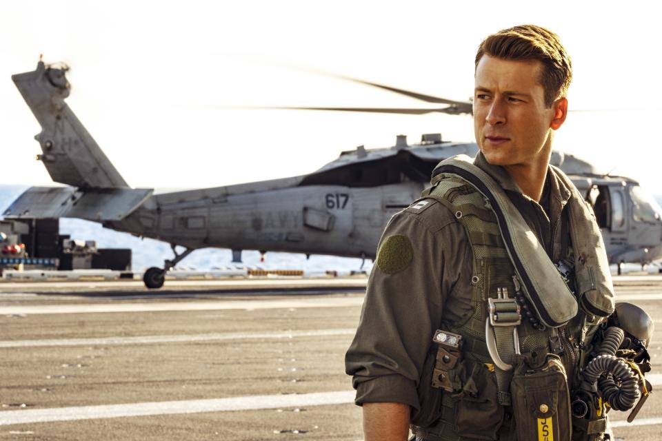 A man in a military uniform stands on an aircraft carrier deck with a helicopter in the background. The scene is from a TV or movie production