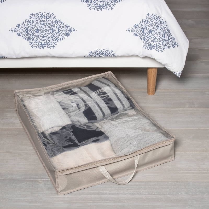 Clear storage box with a variety of folded blankets beside a bed, useful for organizing linens