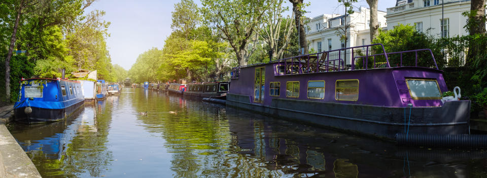 Wide panoramic view of a line of colorful canal boats moored along a lush urban waterway under a clear sky.