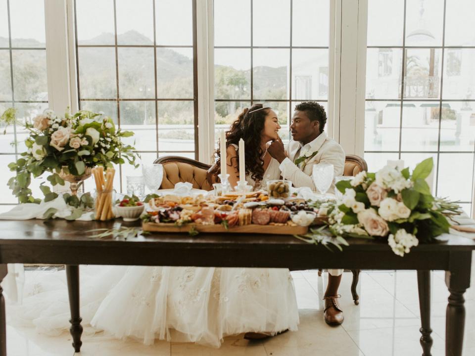 A groom brushes a bride's lip with his thumb as they sit on a couch behind an ornate table.