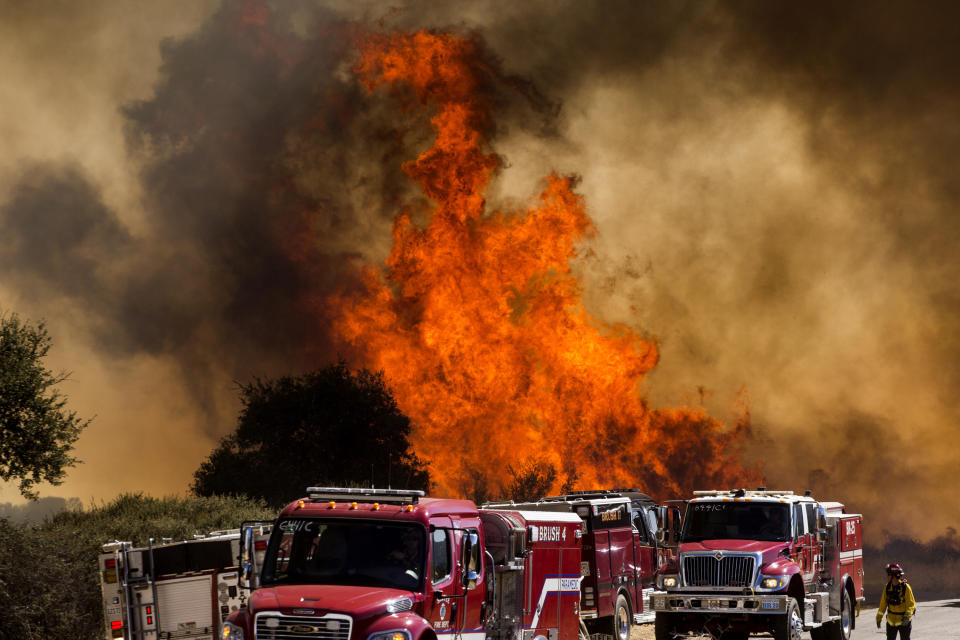 Flames flare behind fire trucks at the Apple Fire in Cherry Valley, Calif., Saturday, Aug. 1, 2020. A wildfire northwest of Palm Springs flared up Saturday afternoon, prompting authorities to issue new evacuation orders as firefighters fought the blaze in triple-degree heat. (AP Photo/Ringo H.W. Chiu)
