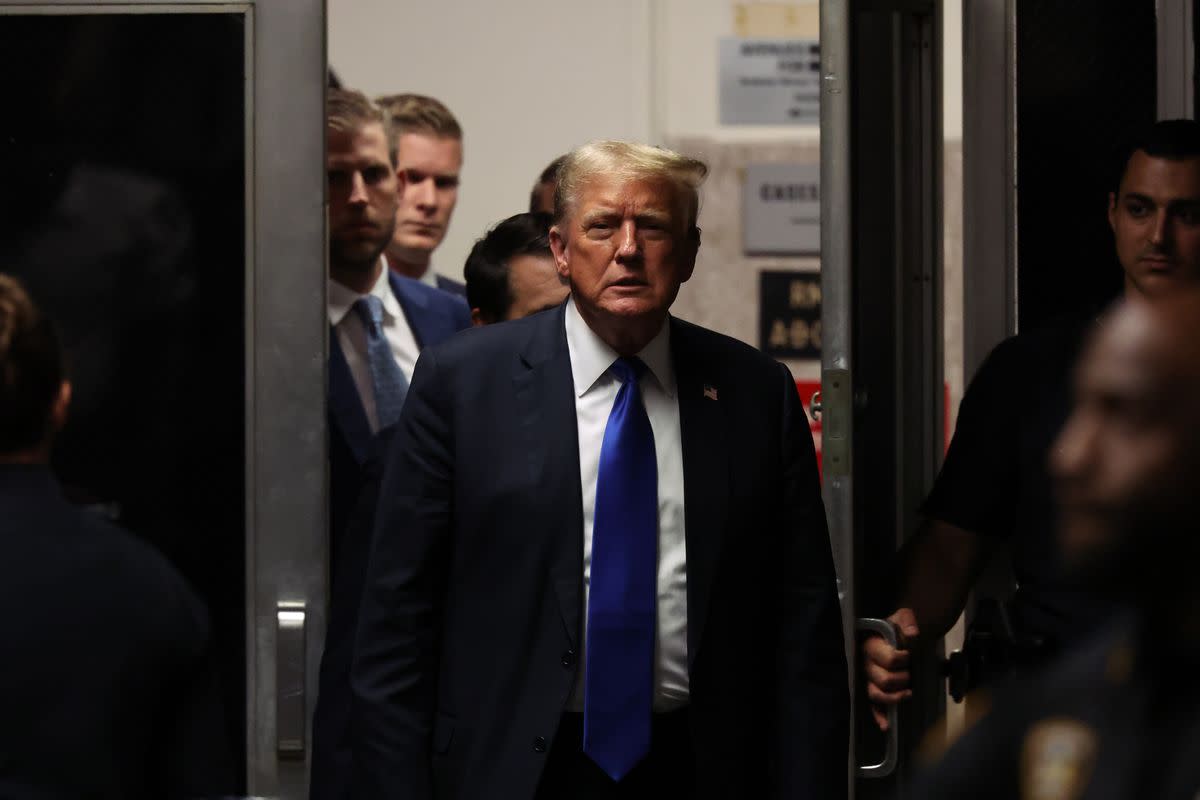 A white man wearing a black suit walks out of a building. Someone holds the door for him. You can see security officers near the front of the photo, and a crowd of people stands behind him. 