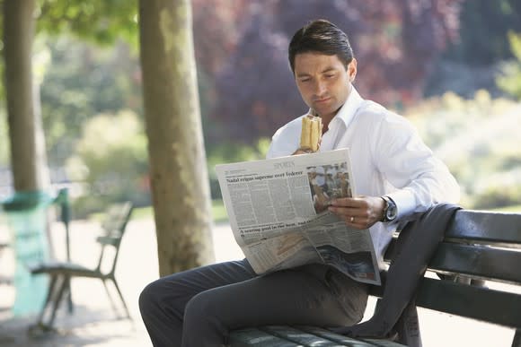 Man in professional clothing reading a newspaper on an outdoor bench while holding a sandwich.