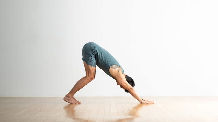 A man in blue shorts and a top practices Downward-Facing Dog with his knees bent. He is on a wood-plank floor with a white wall behind him